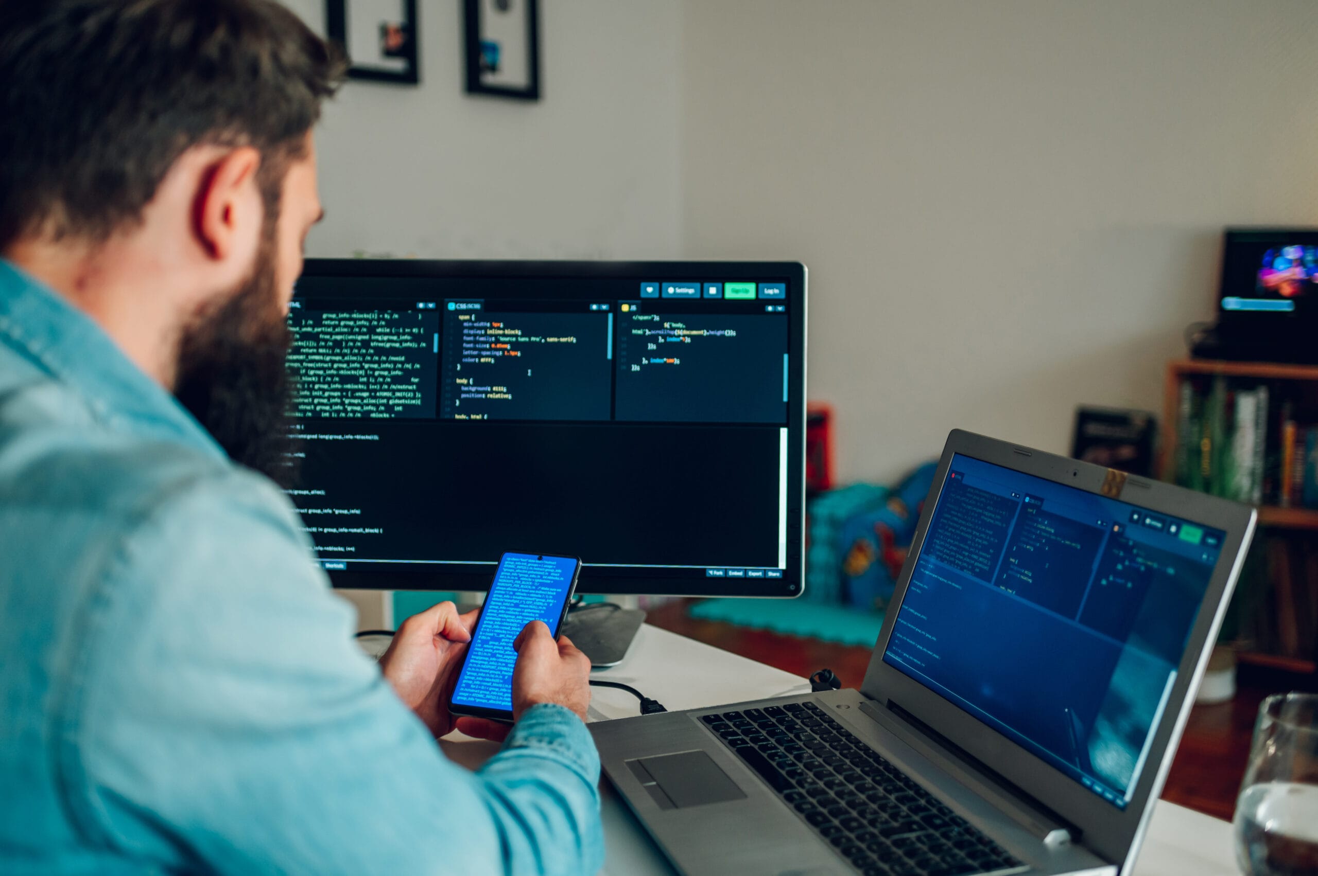 A senior mobile app developer is using his phone to check on code while sitting at his home office surrounded by computer screens. Rear view of an IT expert working on the mobile app.